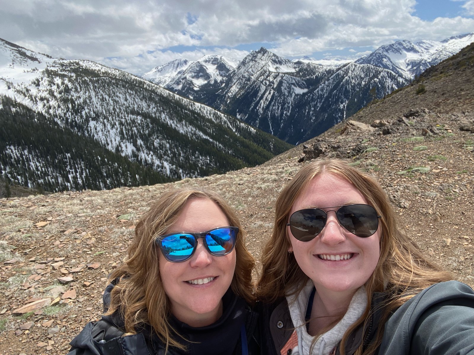 Two people smiling with snow-covered mountains behind them.