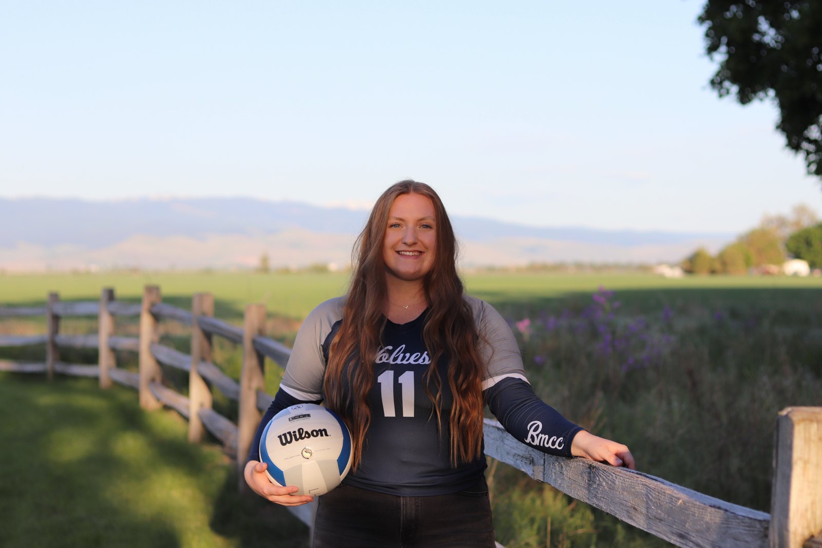 Kenzie in her college volleyball uniform, holding a volleyball beside a fence.